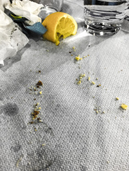 after an eaten up meal on a paper tablecloth you see breadcrumps, a pressed lemon, a glass of water, crunched up servietten and grease stains