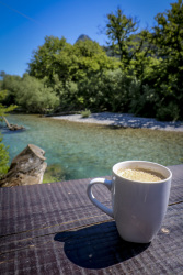 in front of a sunny landscape with a tourquoise river is standing a cup of yummi hot coffee
