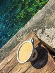 a big white cup with coffee americano is standing on the bank at the tourquise aegean sea