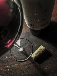 a glass of red wine, a cork and a bottle of wine from puglia on a dark wooden table
