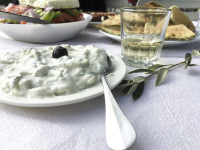 an olive branch next to some plates with tzatziki, greek salad, fried zucchini and a glas of greek white wine