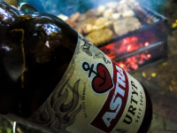 behind a bottle of astra beer there is a glowing grill with much meat on it