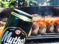behind a can of the greek beer myhtos are a grill full of sausages and steaks