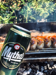 behind a can of the greek beer myhtos are a grill full of sausages and steaks