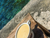 a big white cup with coffee americano is standing on the bank at the tourquise aegean sea