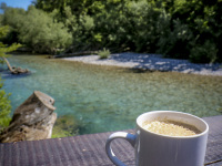 in front of a sunny landscape with a tourquoise river is standing a cup of yummi hot coffee