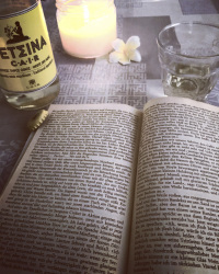 a open site of the book reflections of a marine venus from lawrence durrell, a bottle cair retsina, a glass, a candle and a blossom of frangipani