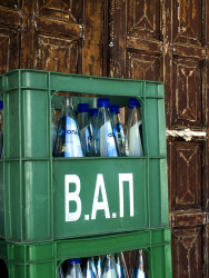in front of a brown wooden door in lindos on rhodes is standing an old green vap drinks crate with blue bottles