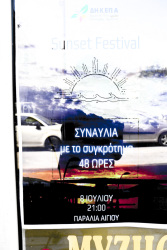 in a glass door of a store in aegion is hanging a plakat for a sunsetfestival, two cars are reflecting in the glassdoor