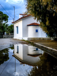 a little white chapel in diakopto reflects in a big puddle in front of it