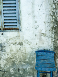 an old grey wall with peeling plaster, with a blue window and a blue greek chair in agios isidoros on rhodes