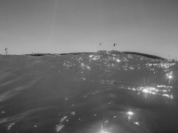 the sun is shining on a wave in prasonisi and in the distance are small kites, black-and-white