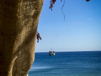 next to a tree trunk you see the blue sea, the blue sky, a boat and the white stone beach in gerolimenas