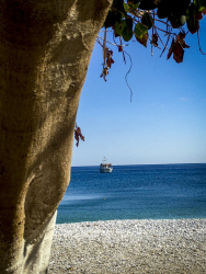 next to a tree trunk you see the blue sea, the blue sky, a boat and the white stone beach in gerolimenas