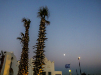 two palms, an oriental house and lanterns are standing in front of a sunset sky with a little white moon at elli beach in rhodes city