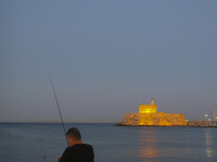 a man is fishing at the mandraki harbor on rhodes with the loveley iluminated fort of st. nikolaos in the background