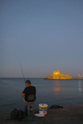 a man is fishing at the mandraki harbor on rhodes with the loveley iluminated fort of st. nikolaos in the background