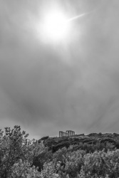 the sun peeks through clowds above the columns of kap sounion, black-and-white